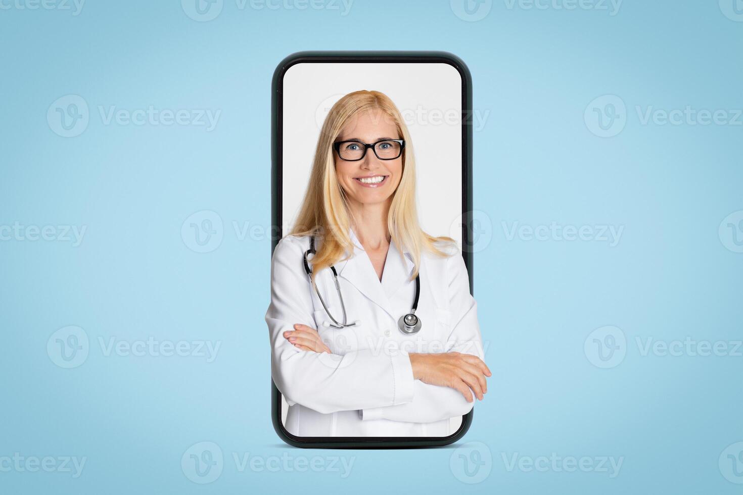 A smiling lady doctor wearing a white coat and stethoscope is visible on a smartphone screen against a blue background. She is looking directly at the camera with her arms crossed. photo