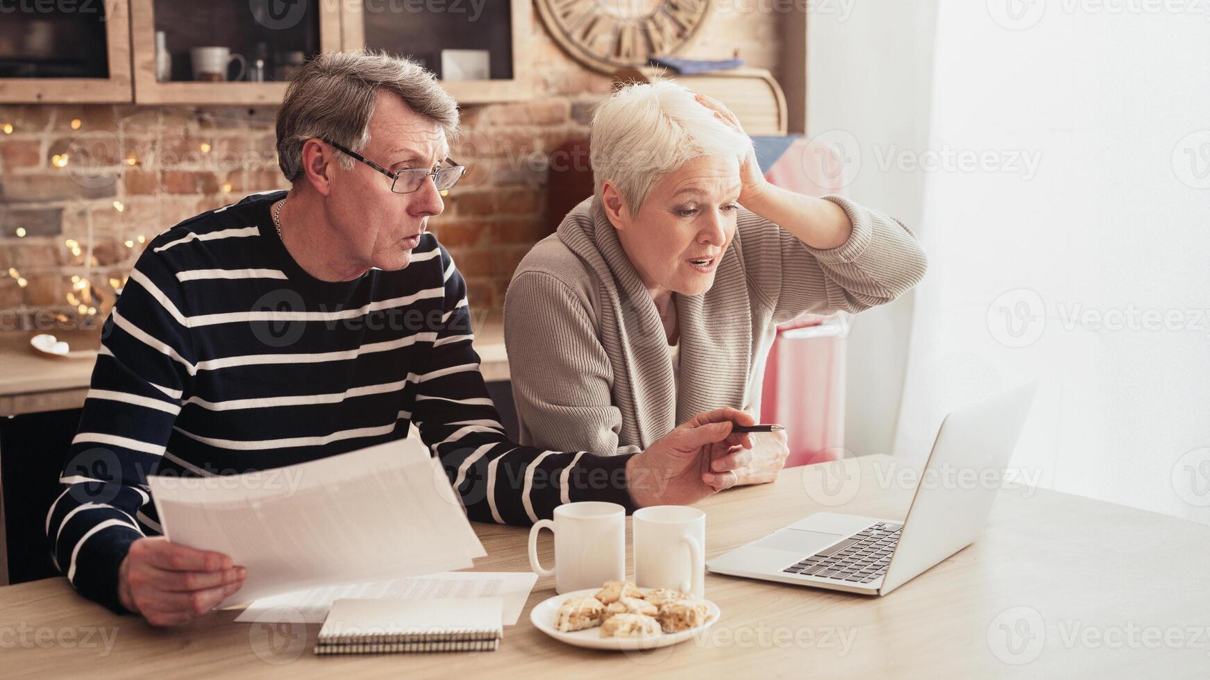 This image shows a senior couple sitting at a kitchen table, reviewing bills and a laptop together. They both have concerned expressions on faces, suggesting they are facing a financial challenge photo