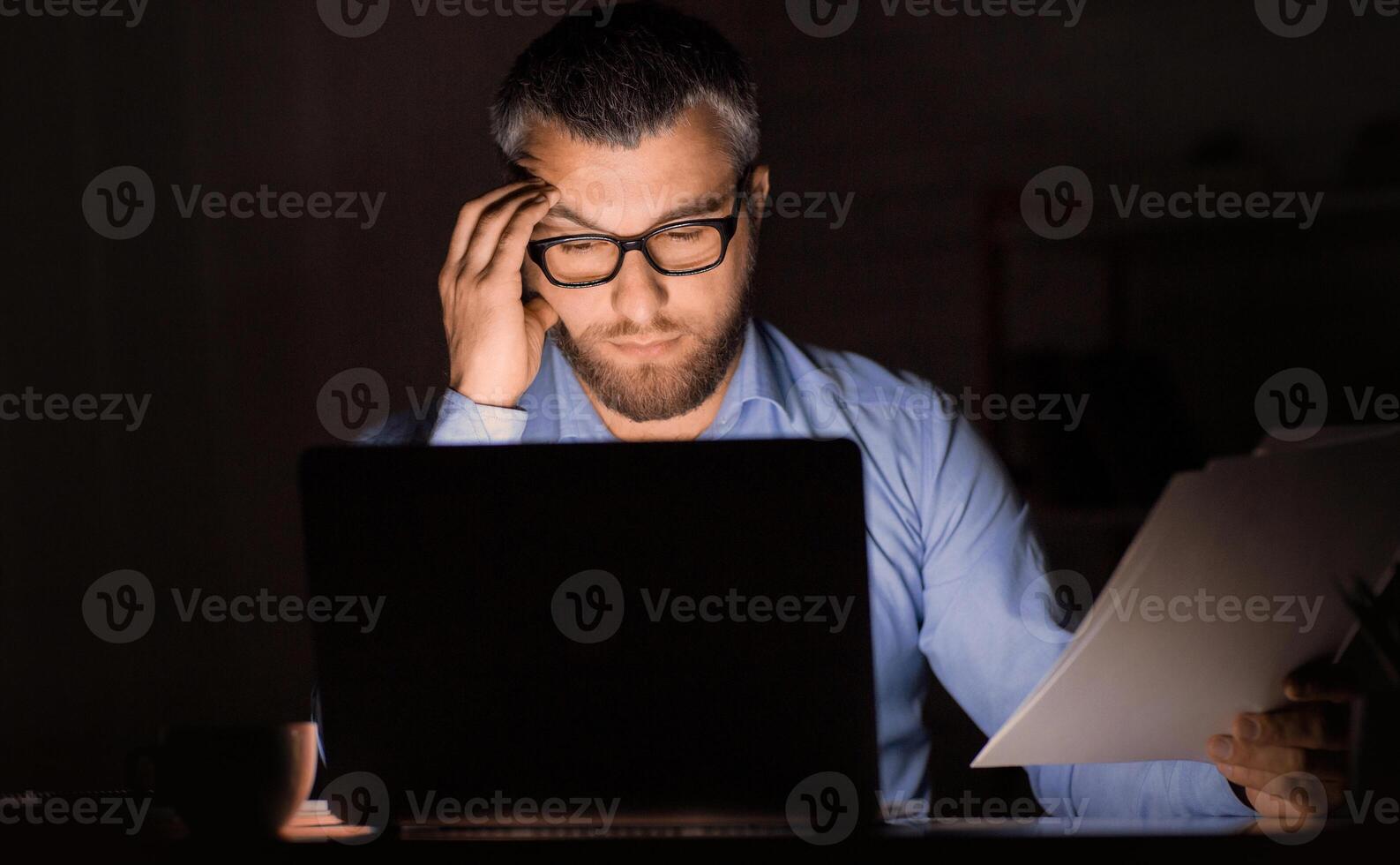 A man in glasses is seen working late in a dimly lit office. He holds documents in one hand while looking intently at a laptop screen. The setting suggests diligent effort and focus photo