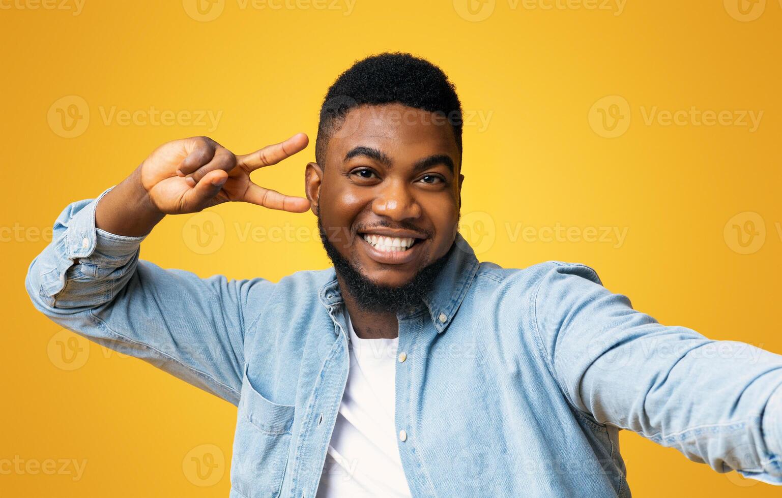 Say Cheese. Joyful black millennial guy showing peace sign while taking selfie, yellow studio background with free space photo