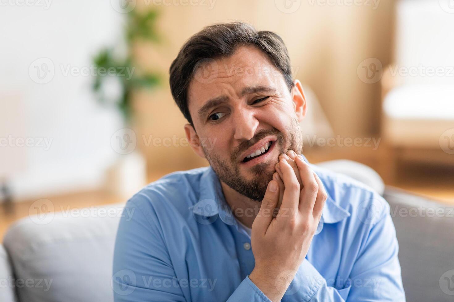 A man in discomfort is seated on a couch, holding his face in pain due to a toothache. His expression reveals his suffering as he tries to alleviate the ache. photo
