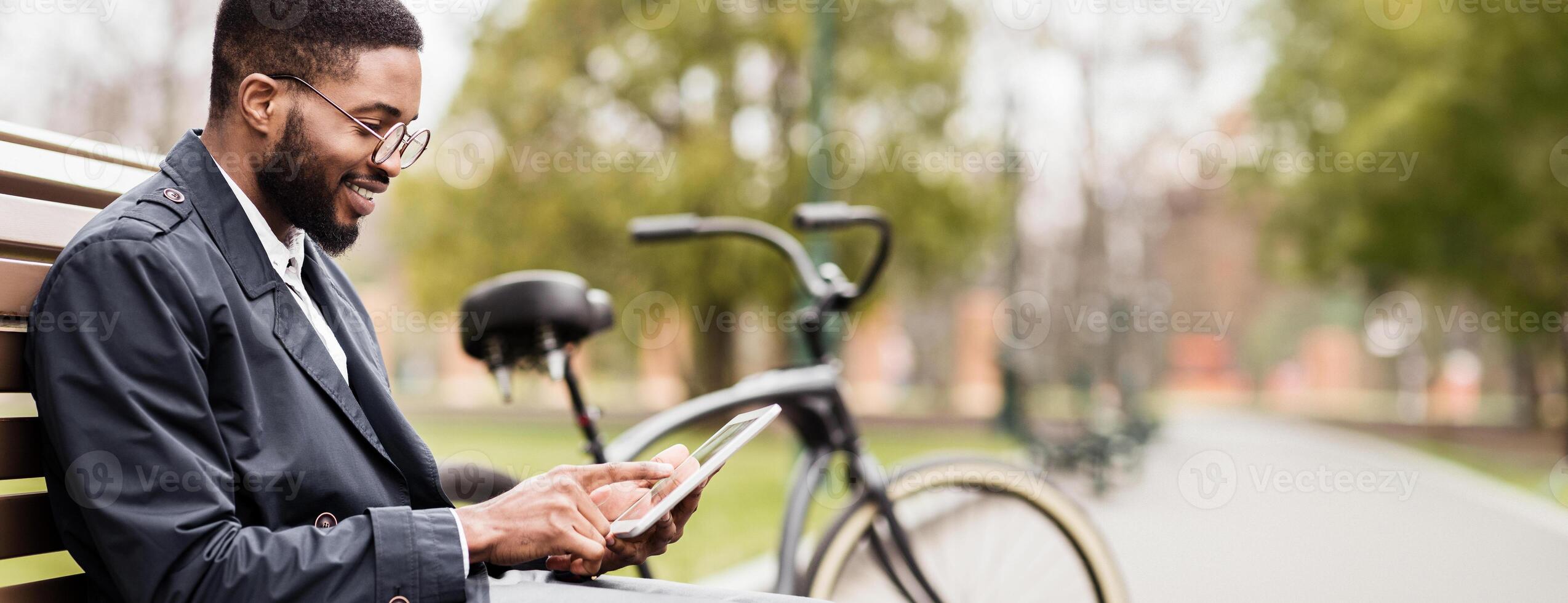 A professional black man is in a park, seated on a bench with a tablet and bicycle. The scene is relaxed and contemporary, capturing a moment of productivity in a natural setting. photo
