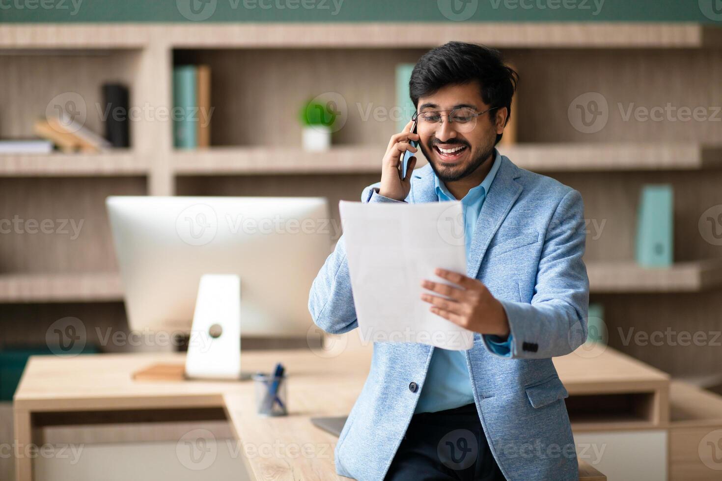 A smiling Indian man in a blue blazer is talking on the phone while reading documents. He is standing at a desk in a modern office. photo