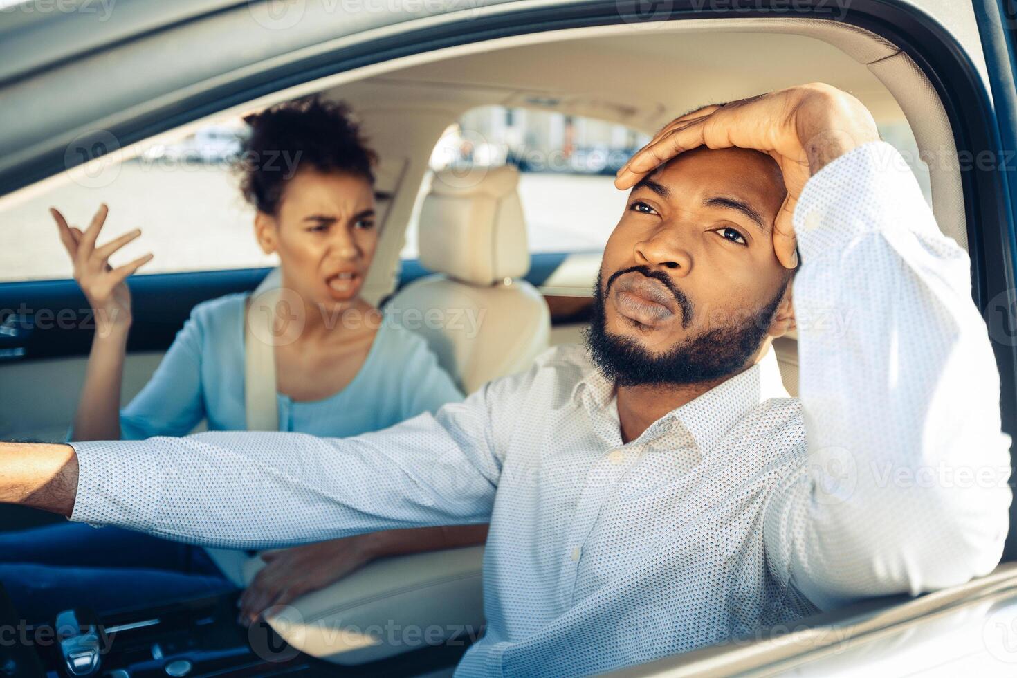 Black man driving a car appears stressed and frustrated as a woman in the passenger seat yells at him. He has his hand on his forehead as he stares forward while she gestures with her hand raised. photo