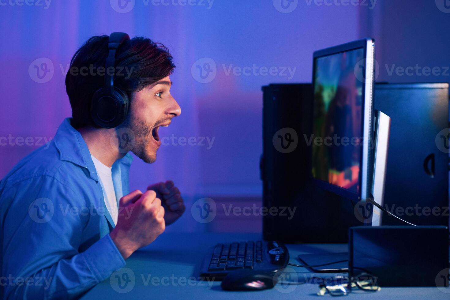 A young man wearing headphones sits at his computer, excitedly playing a game. His mouth is open in surprise and his fists are clenched in victory as he watches the action unfold on the screen. photo