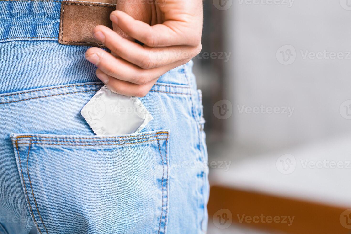 A close-up shot of a persons hand placing a condom into the back pocket of their blue jeans. The hand is reaching into the pocket, and the condom is visible as it is being inserted, cropped photo