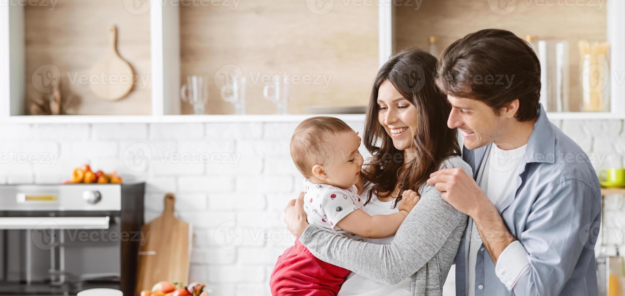 This image shows a happy family of three in their kitchen. The parents are smiling and holding their baby in their arms. The baby is looking up at the parents and appears to be happy. photo