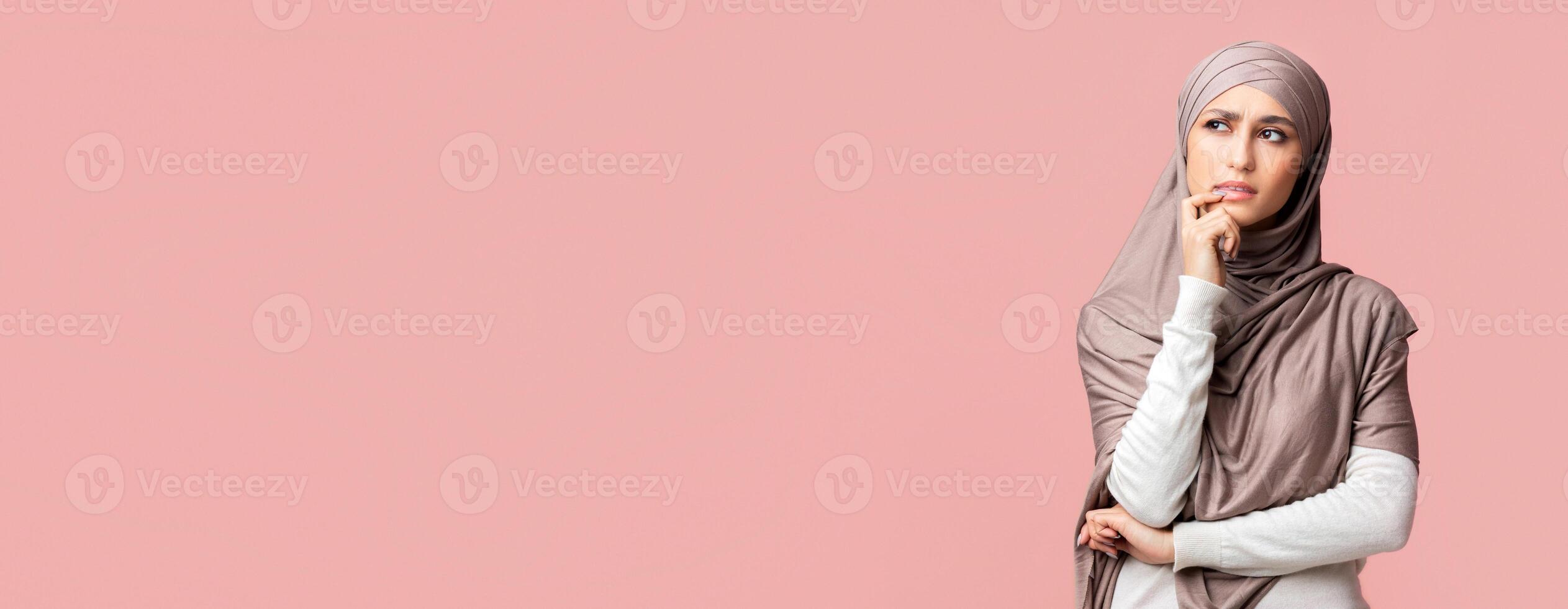 Pensive arabic woman in hijab thinking about something and looking away on pink studio background, panorama with free space photo