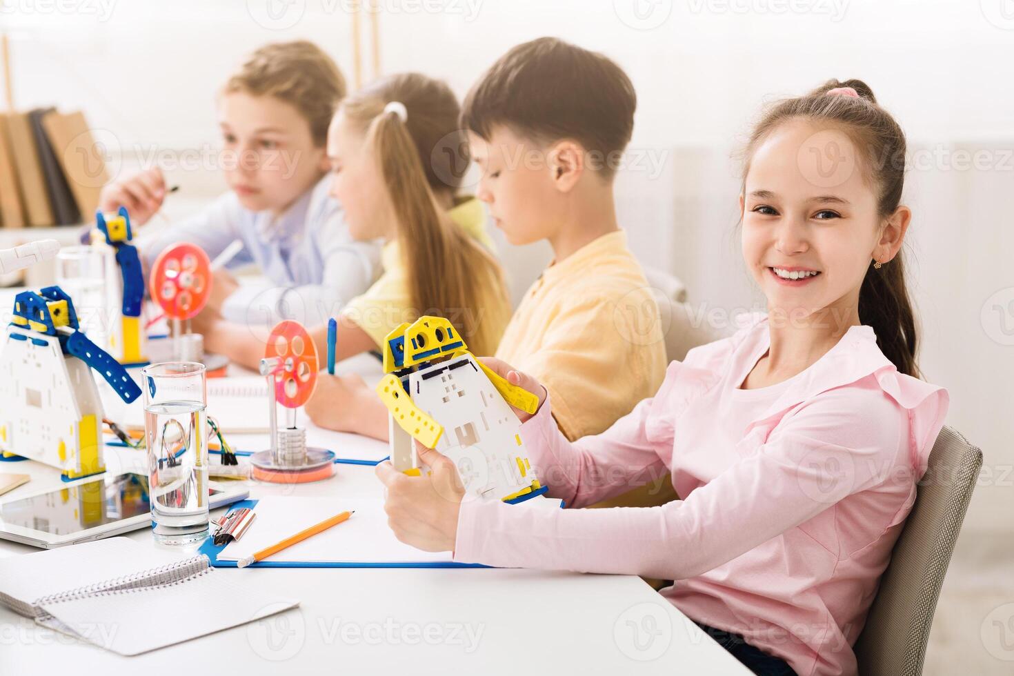 This image shows a group of children in a classroom setting, engaged in a STEM activity. They are working on building robots, and the girl in the foreground is holding up her finished creation photo