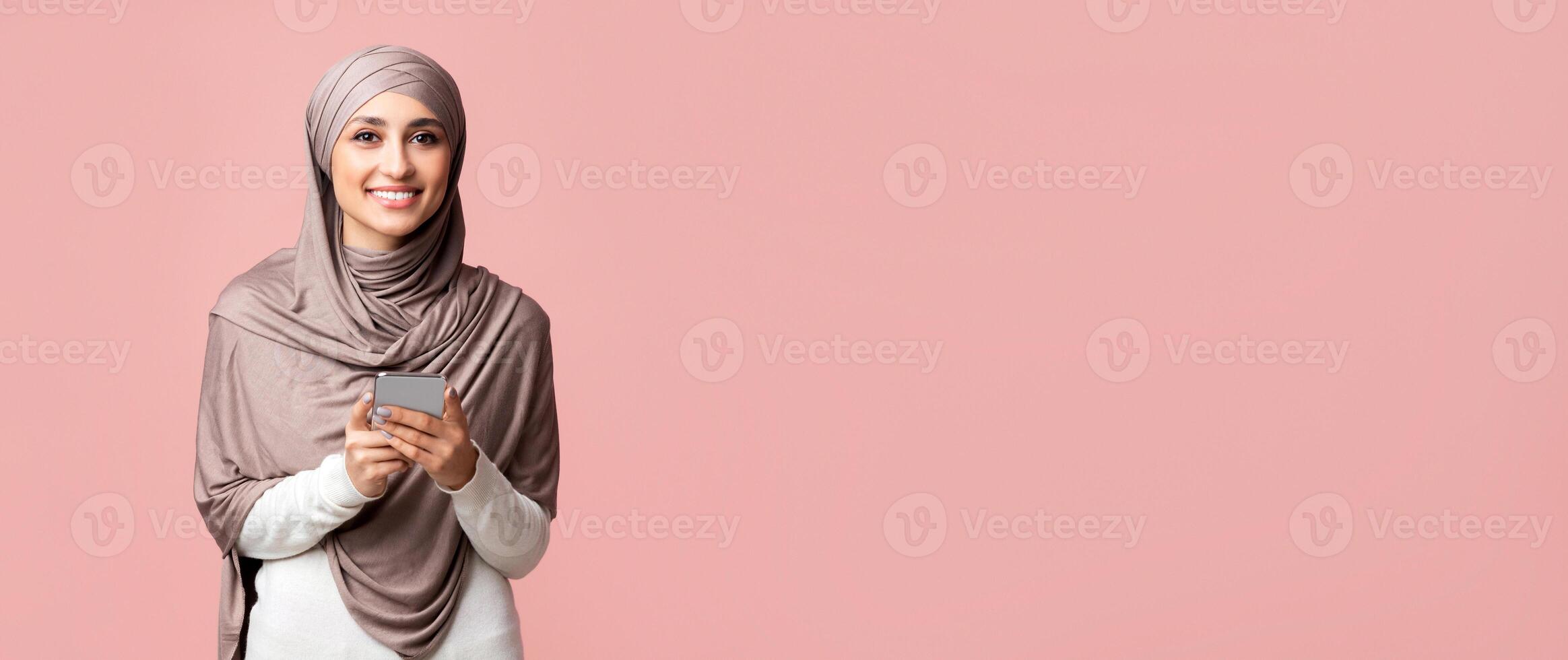 Best Apps for Muslims Concept. Smiling islamic girl in headscarf holding smartphone and looking at camera over pink studio background, free space photo
