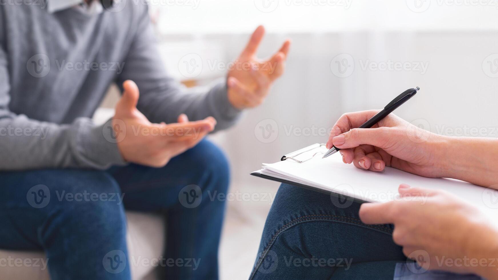 A person is seated in a neutral-colored office space, engaging in a discussion with another individual who is taking notes on a pad, cropped photo
