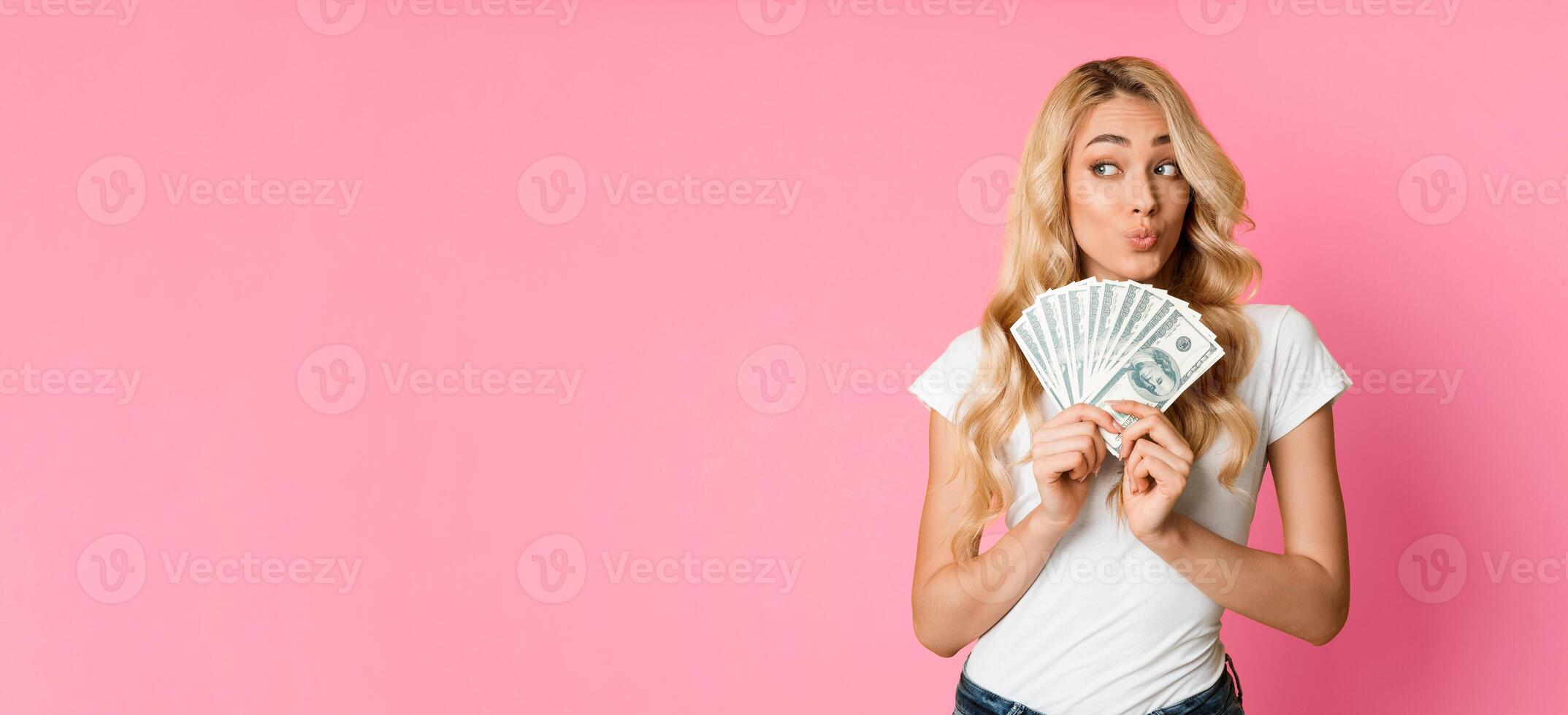 A young woman with long blonde hair is holding a fan of cash in front of her face. She is smiling and looking to the side. The background is a bright pink, panorama with copy space photo