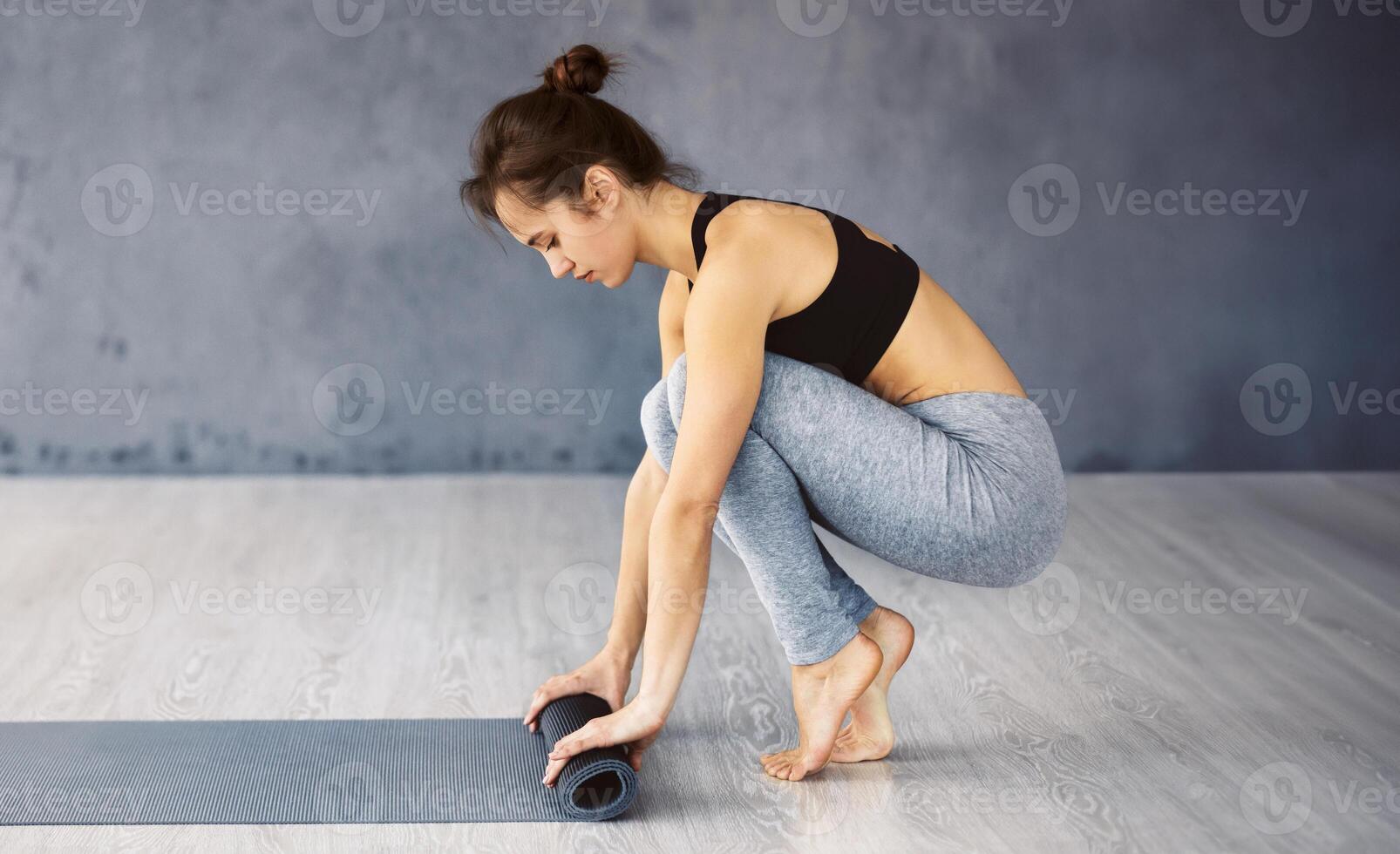 A focused young woman is in the process of unrolling her yoga mat on a wooden floor in a tranquil studio setting, preparing for her exercise routine. photo