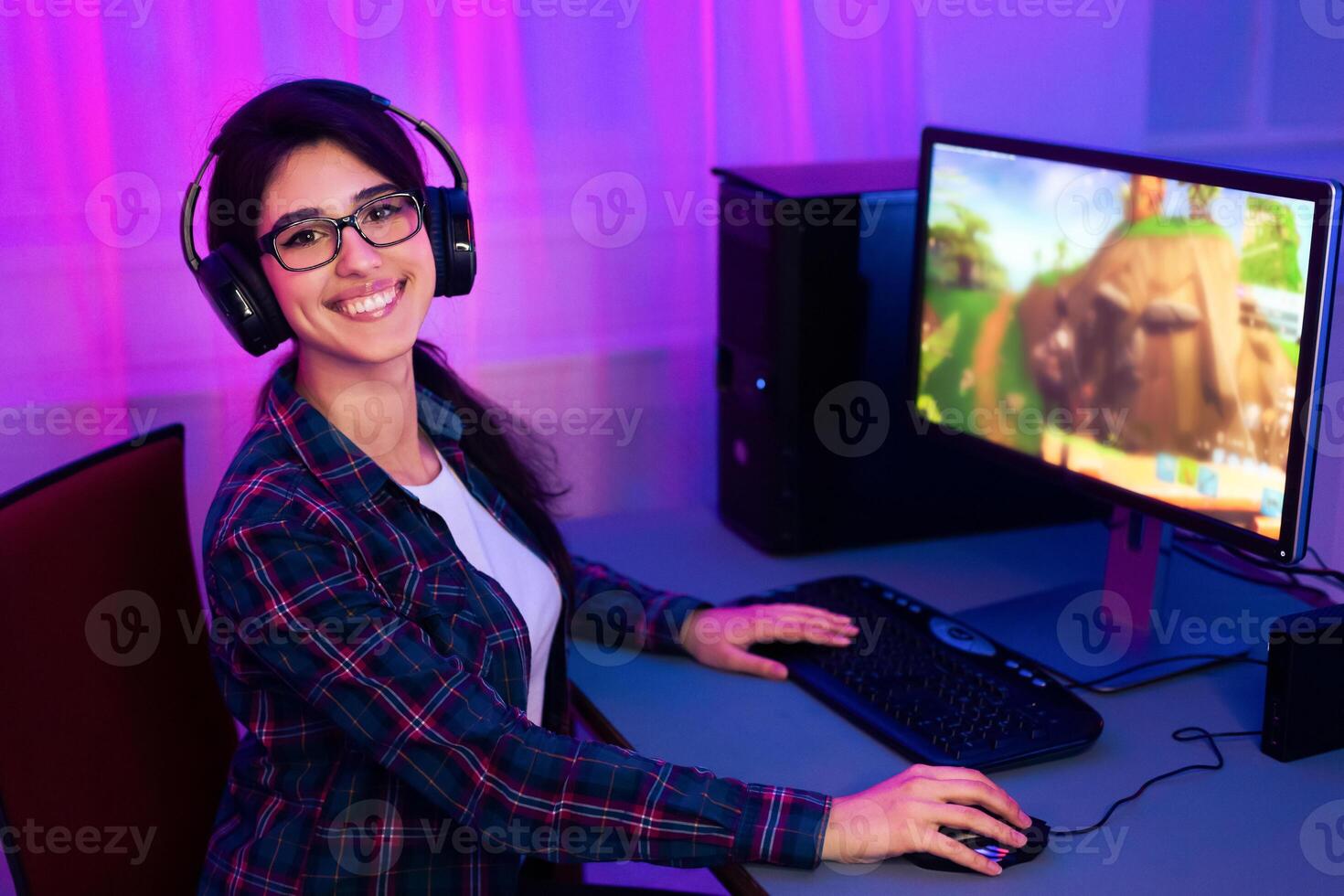 A young woman with long dark hair is smiling as she plays a computer game. She is wearing a plaid shirt and headphones. The computer monitor in the background shows an animated game. photo