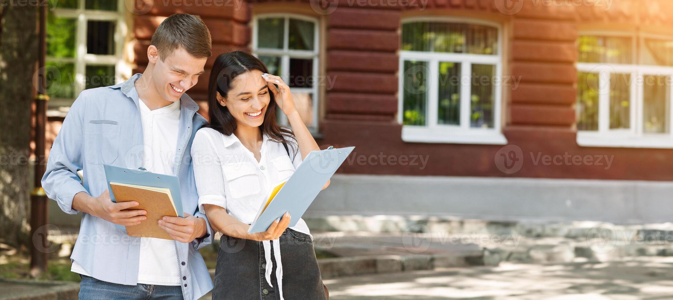 Portrait of cheerful couple of university friends laughing outdoors while checking classes schedule, reading information in notepad photo