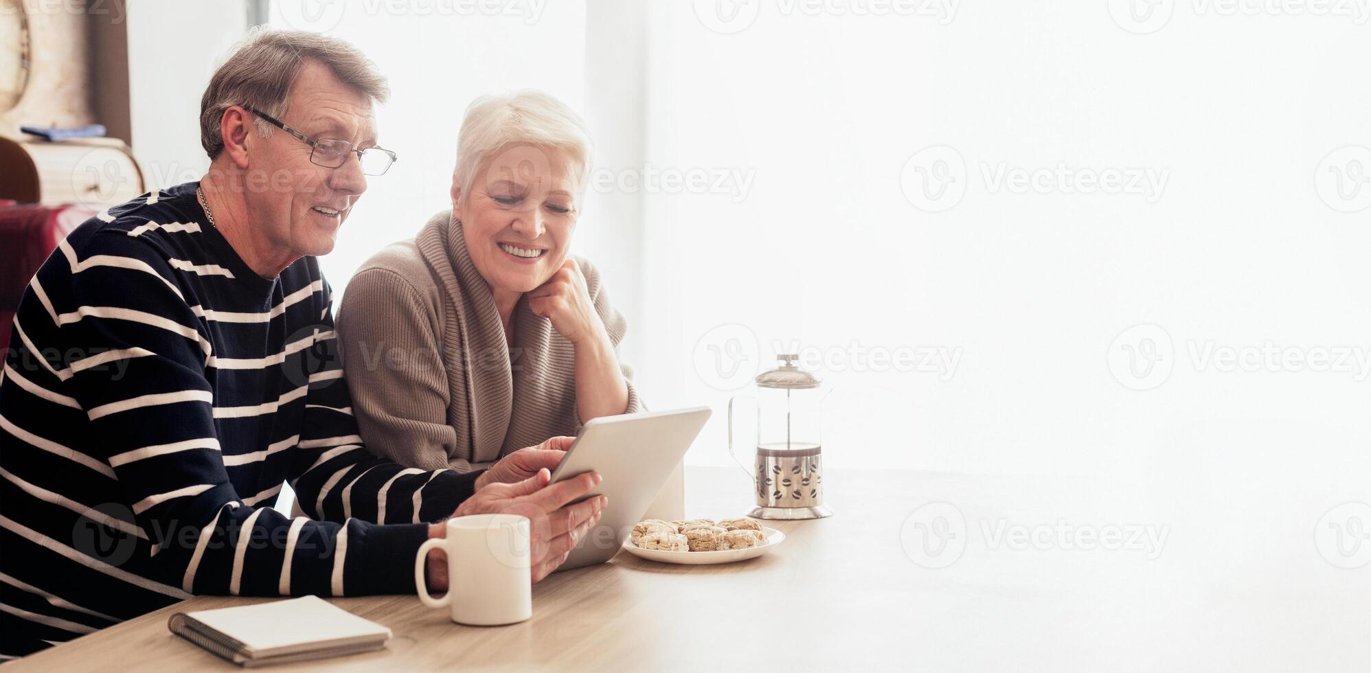 A happy senior couple sits at a table in their home, browsing the internet on a tablet. The man is wearing a striped sweater and glasses, while the woman is wearing a beige sweater and smiling. photo