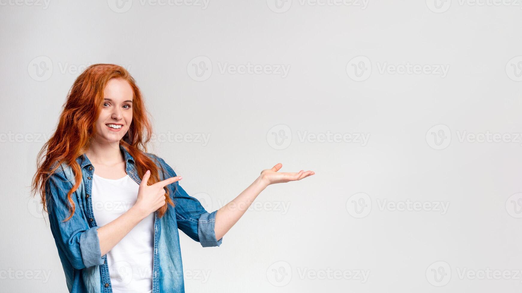 A cheerful redhead woman wearing a denim shirt over a white top is standing against a plain light background. She is looking at the camera with a smile while making a presenting gesture, copy space photo