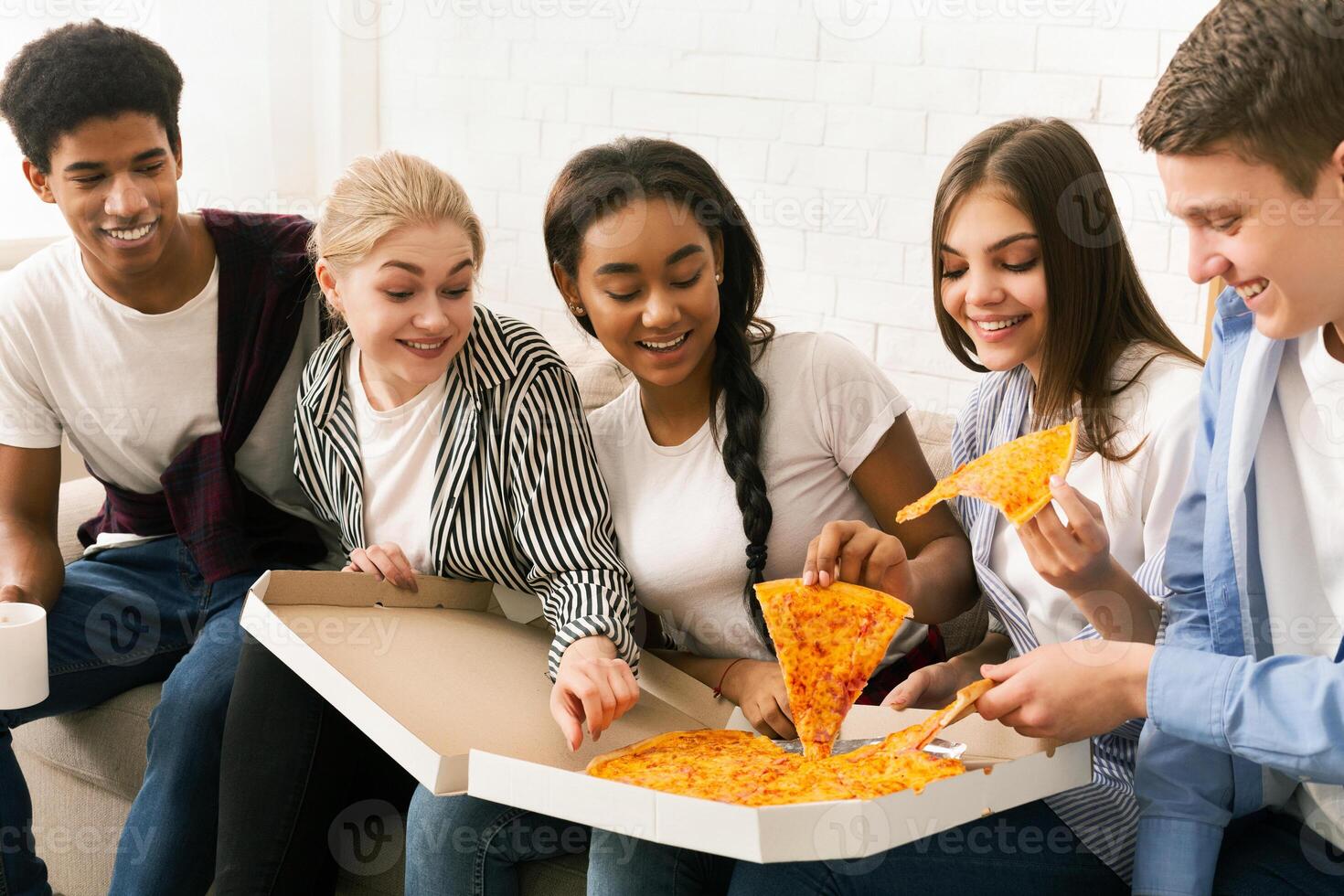 A group of friends sitting on a couch, enjoying slices of pizza. The diverse group is engaging in casual conversation while savoring their meal together. photo