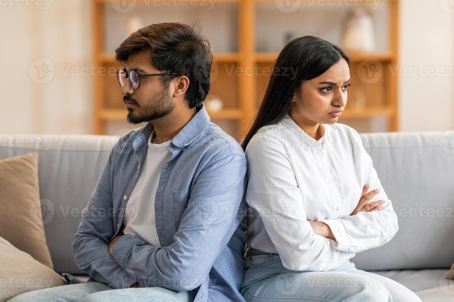 Indian young couple sits back-to-back on a couch, both with arms crossed, appearing to be in the midst of an argument in a bright living room. photo