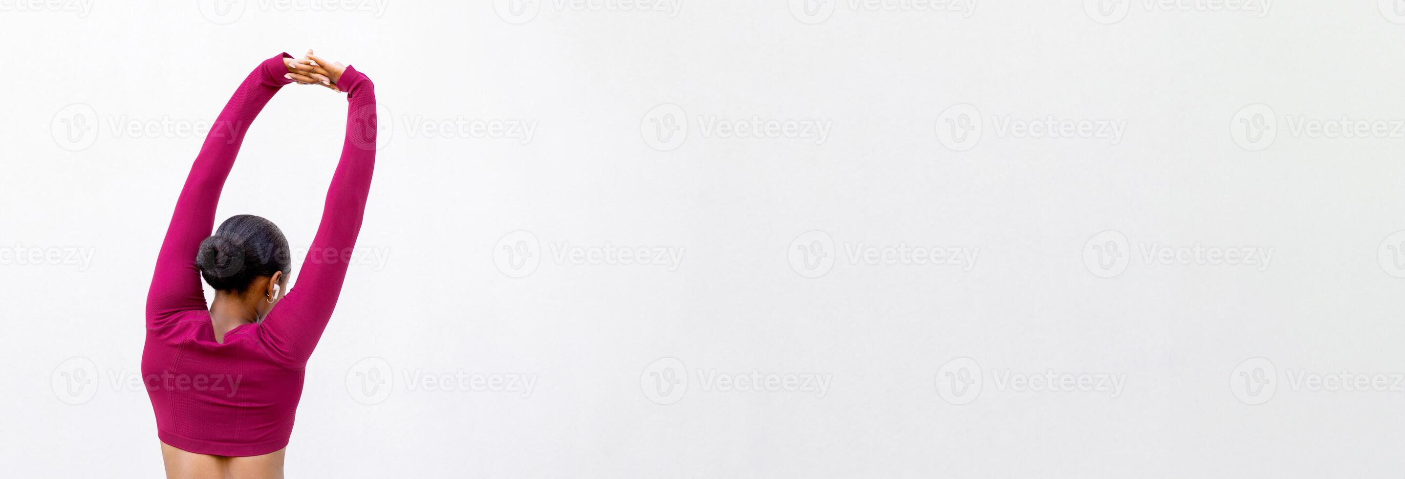 Rear view of athletic black lady with beautiful well-fit body wearing pink tight sportswear stretching arms on white blank wall background, panorama with copy space. Fitness, pilates, workout, sport photo