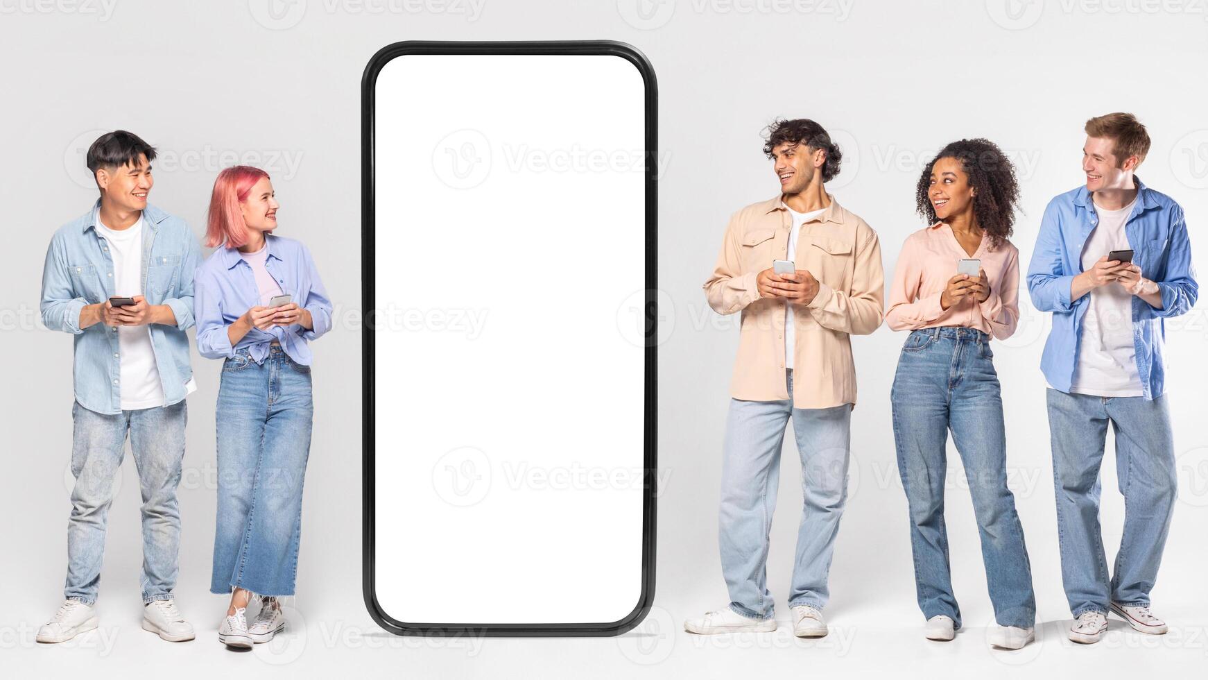 Multiracial people stand in front of a giant phone mockup with blank white screen. They are all looking at the phone and holding their own smartphones. photo
