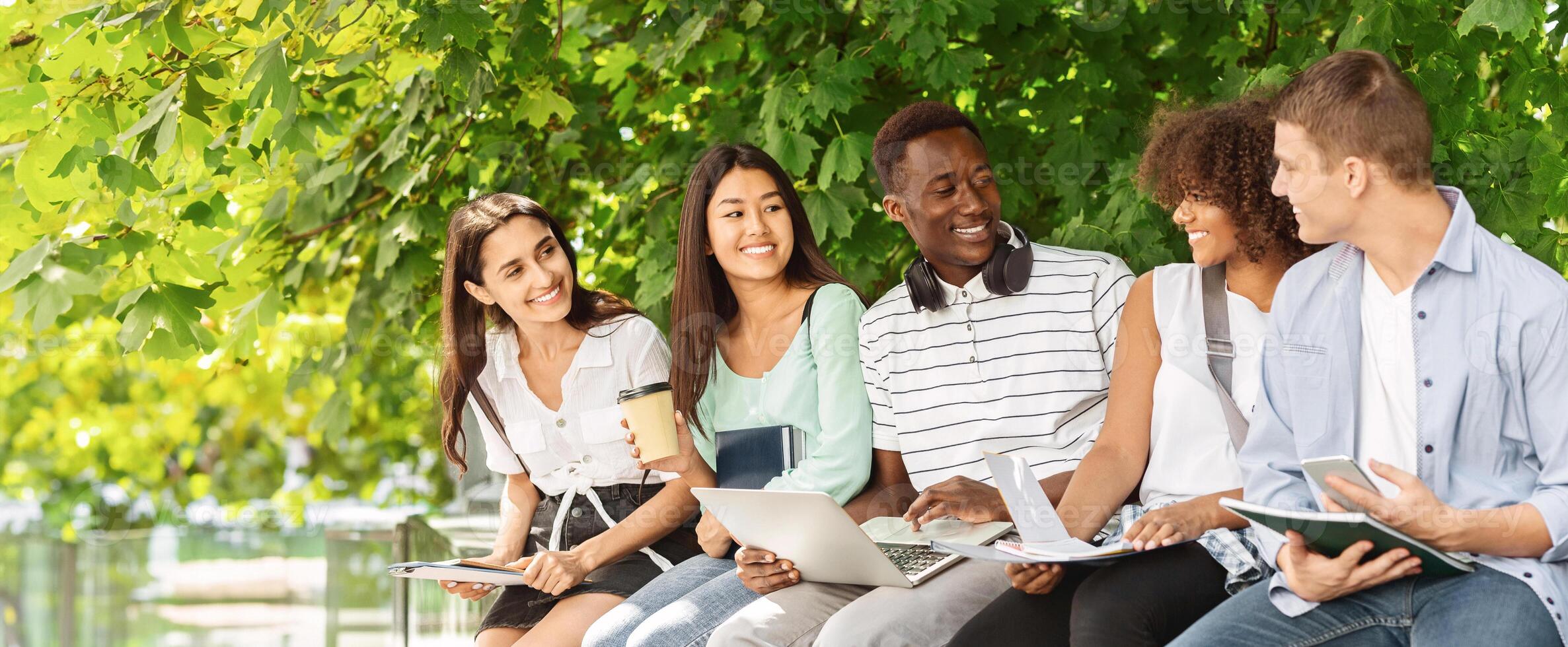 Group of multicultural students sitting in the courtyard of university, chatting and studying together, preparing for lessons photo