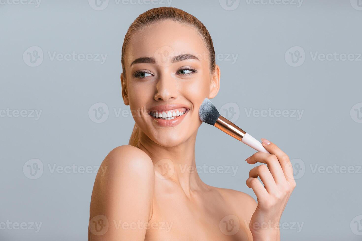 A woman with blonde hair smiles as she applies makeup to her face with a brush. She is wearing no clothing and the background is a simple gray color photo