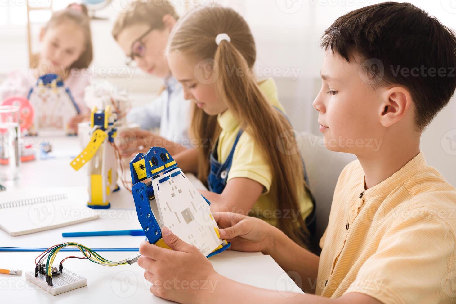 A young boy sits at a table in a classroom and builds a robot with other children. The boy is concentrating on his work as he holds a piece of the robot and examines it. photo