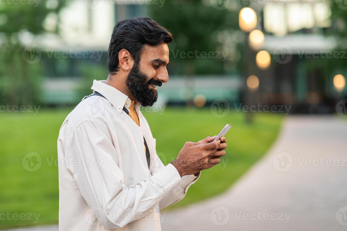 A bearded Indian man is focused on his cell phone screen, possibly reading or texting. His attention is completely captured by the device as he stands still. photo