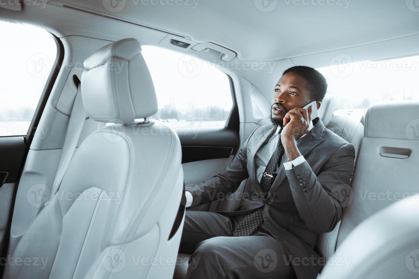 African American businessman in a grey suit is sitting in the backseat of a car, looking out the window while speaking on his phone. The car has a light-colored leather interior. photo