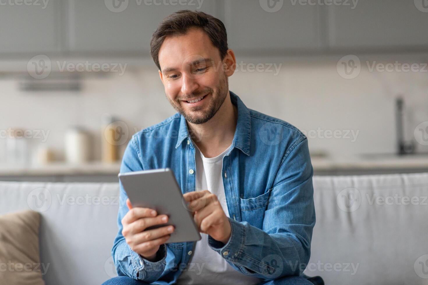 A man, wearing a blue denim shirt, sits on a white couch in a living room setting. He holds a tablet in his hands and scrolls through it with a smile on his face. photo