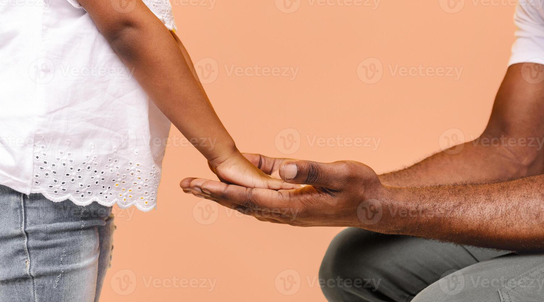 A little girl with brown skin reaches out to an adult males open hand, extending her hand towards him. The man is seated, wearing grey pants and a white shirt, cropped photo
