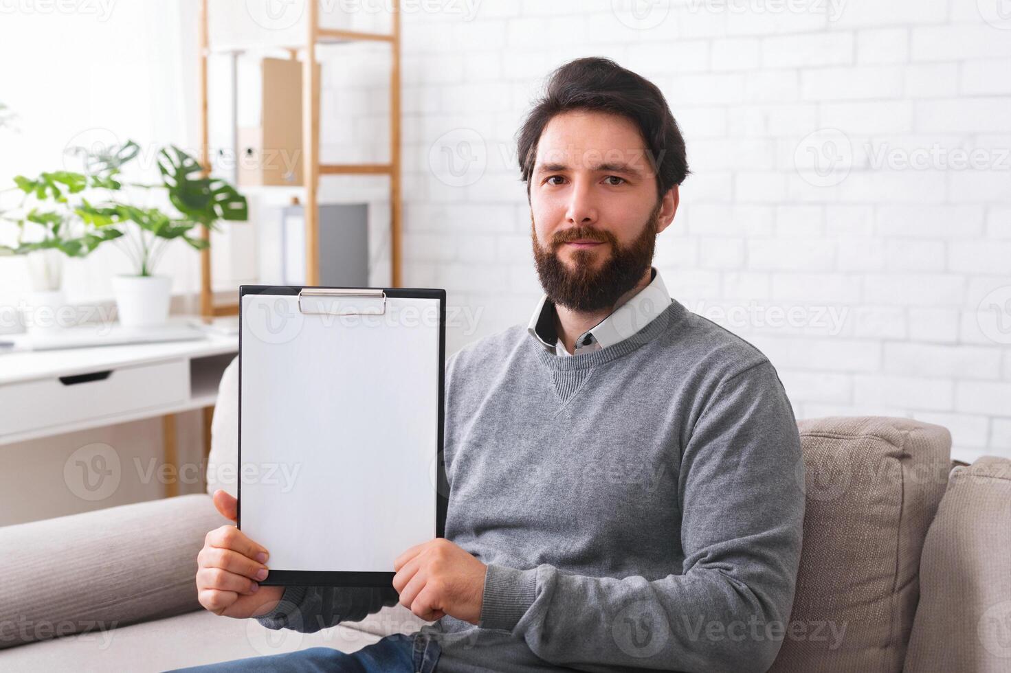 Confident psychotherapist showing white blank clipboard at camera, sitting in office, free space for announcement photo