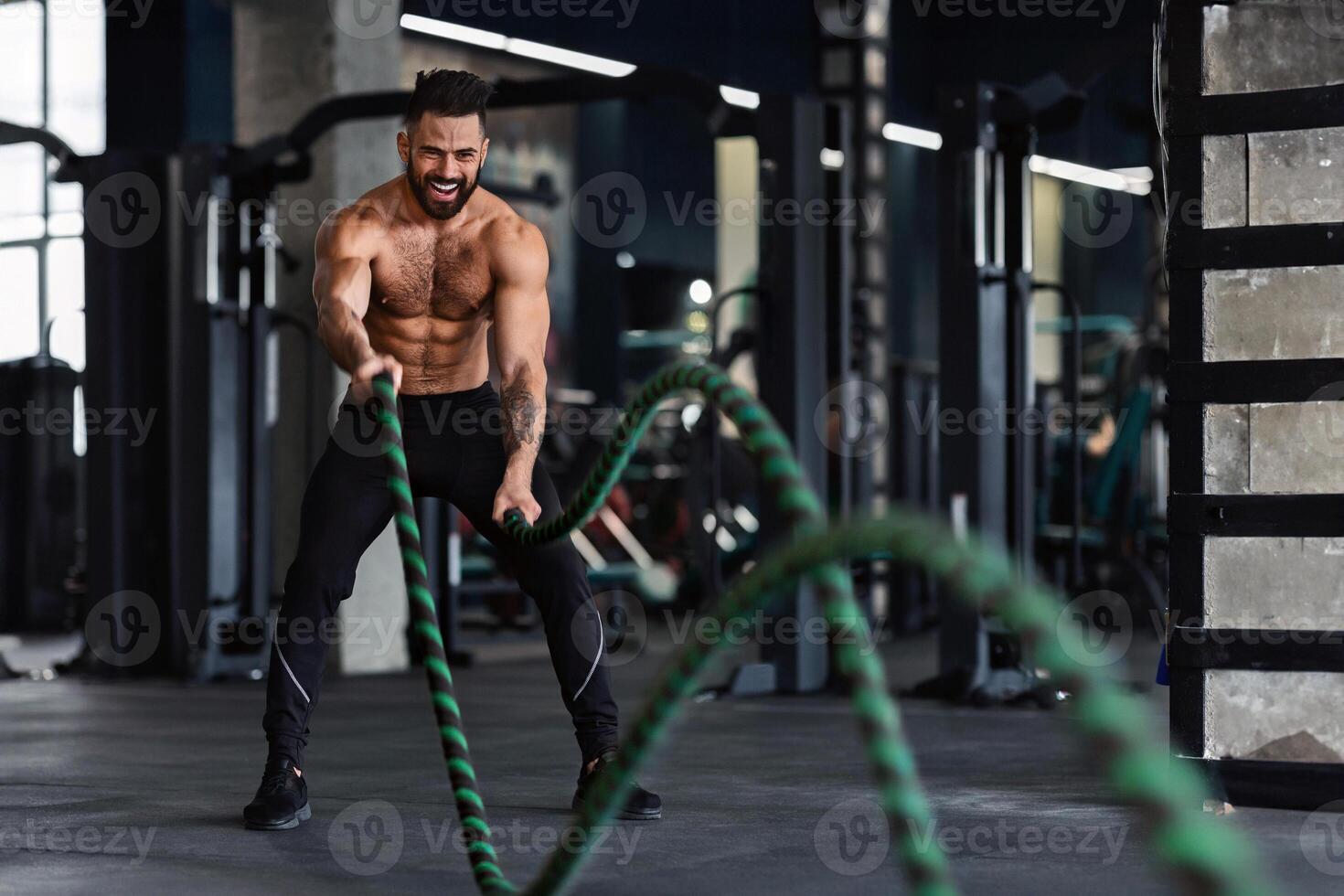 Crossfit training. Naked muscular man exercising with battle ropes at gym, free space photo