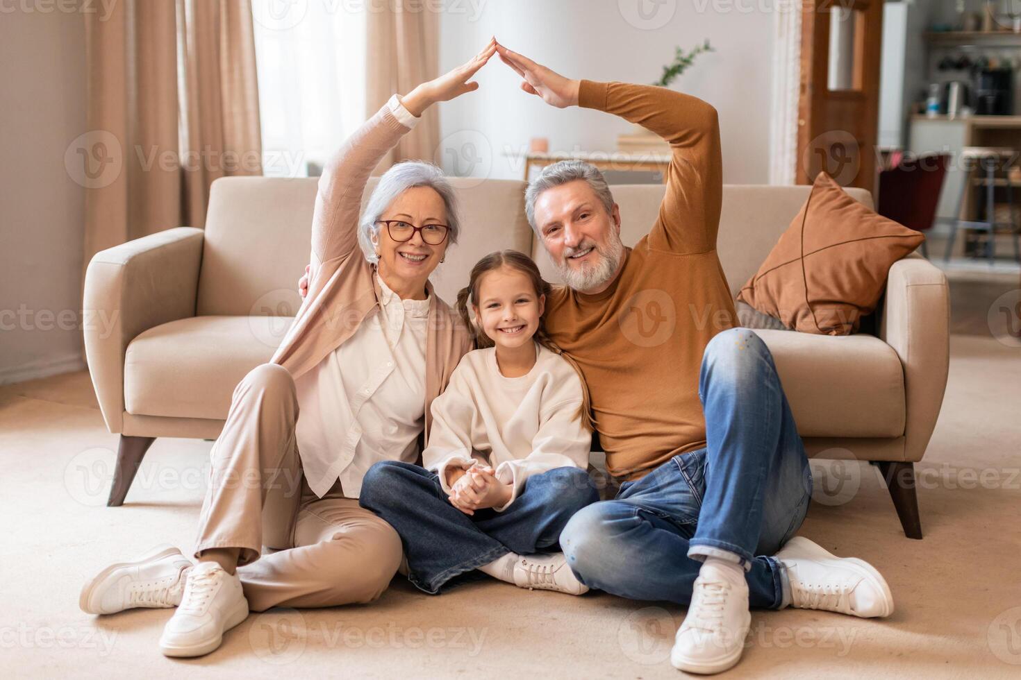 A smiling elderly couple and a young girl sit on the floor in a cozy living room, forming a house outline with their hands above them, symbolizing unity and family love. photo