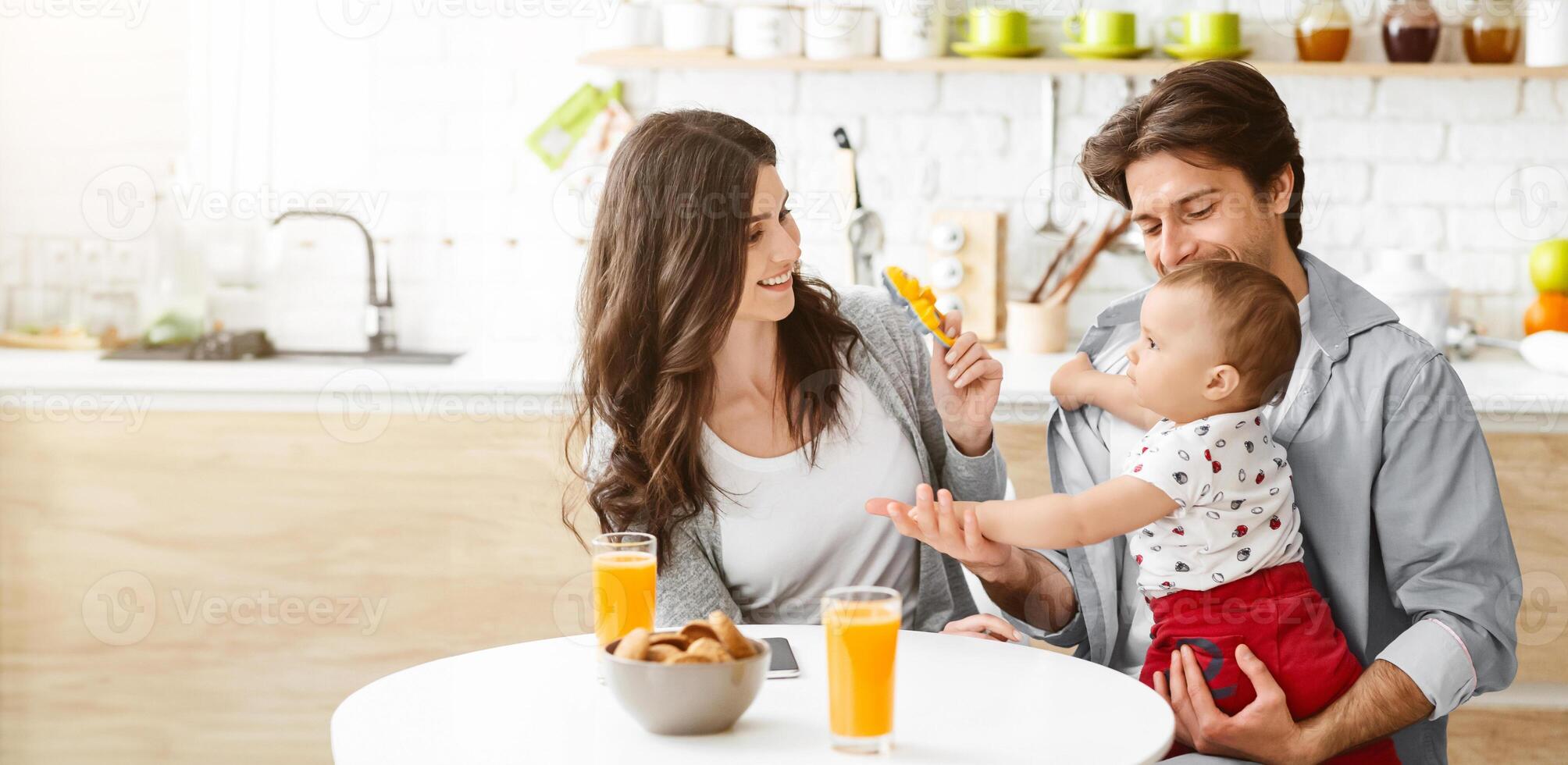 A happy family enjoys a meal together in their kitchen. The father holds their baby, who is reaching out to the mother who sits across the table, holding a yellow toy in her hand, copy space photo