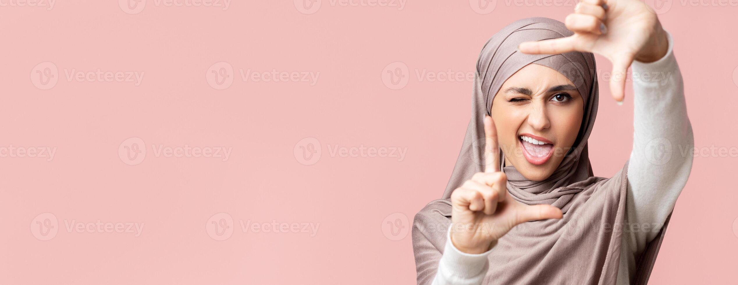 Playful islamic girl in hijab framing face with fingers and winking, pretending to capture photo, pink studio background with free space, closeup photo