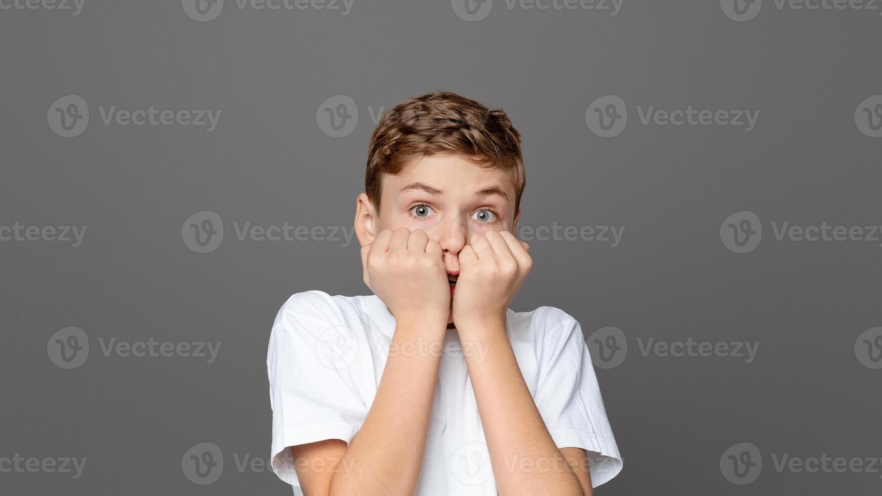 Teenager boy biting his nails in a pose reflecting anxiety and fear, isolated on a grey backdrop, capturing a common youngster behavior photo