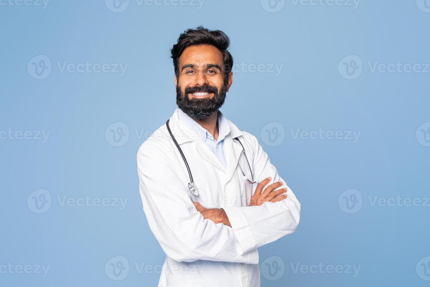 A confident Indian doctor is smiling warmly, dressed in a white lab coat with a stethoscope draped around his neck, standing against a plain blue background. photo