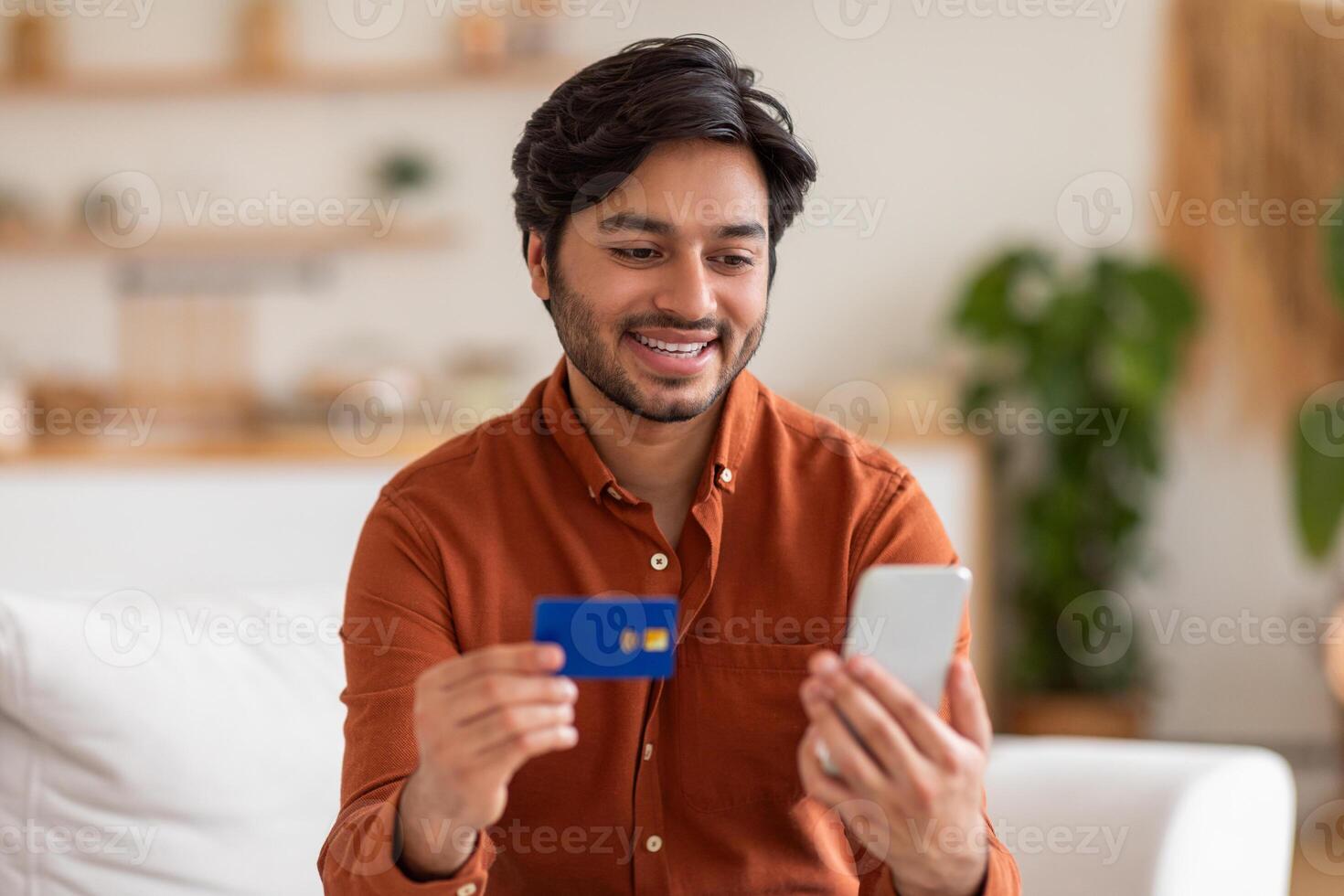 A young Arab man, wearing a rust-colored button-down shirt, sits on a white couch in a home. He holds a credit card in his left hand and a white smartphone in his right hand. photo