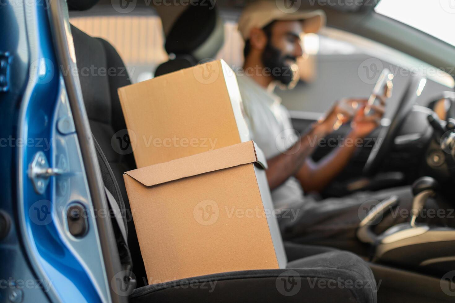 Indian delivery driver is sitting in his parked car, checking his smartphone. Two cardboard boxes are placed on the passenger seat, indicating he is likely in the middle of his delivery route photo
