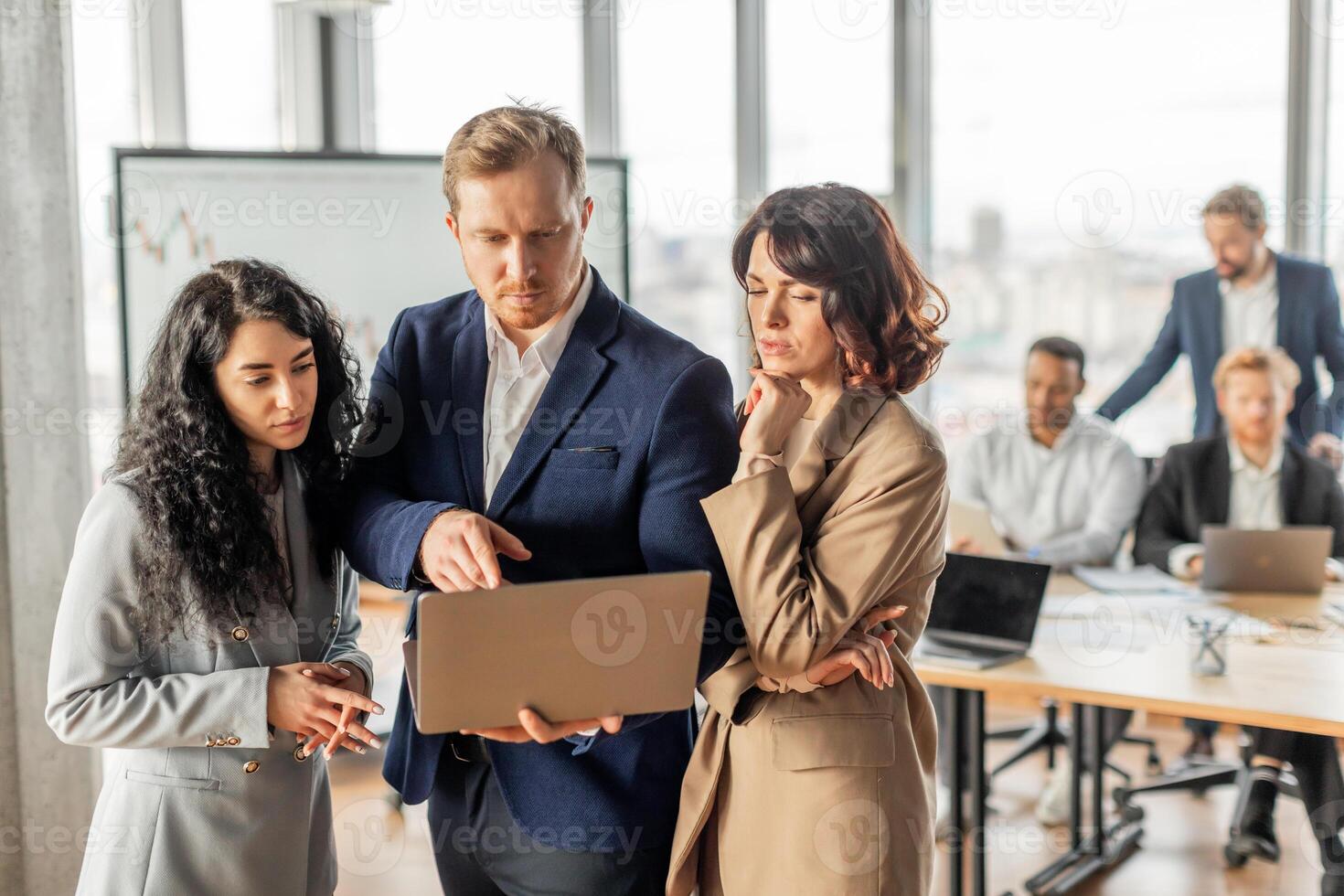 A group of business professionals are gathered around a table in a modern office setting. One man holds a laptop open, pointing at the screen while talking to women standing beside him. photo