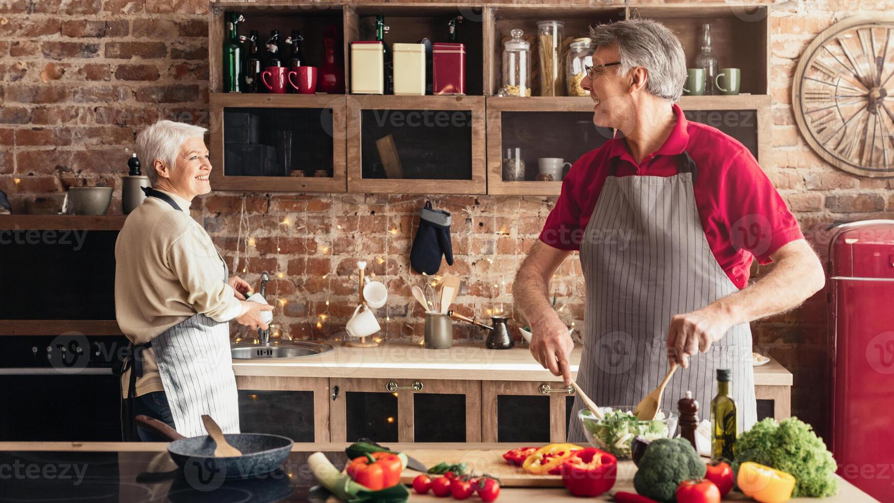 Joyful elderly husband and wife preparing dinner together at modern kitchen. Man making vegetable salad, woman washing dishes photo