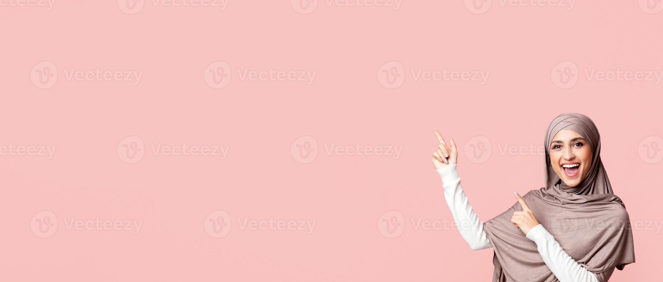 Great offer. Cheerful arabic girl in hijab pointing at copy space on pink background with two hands, showing or advising something, panorama with empty space photo