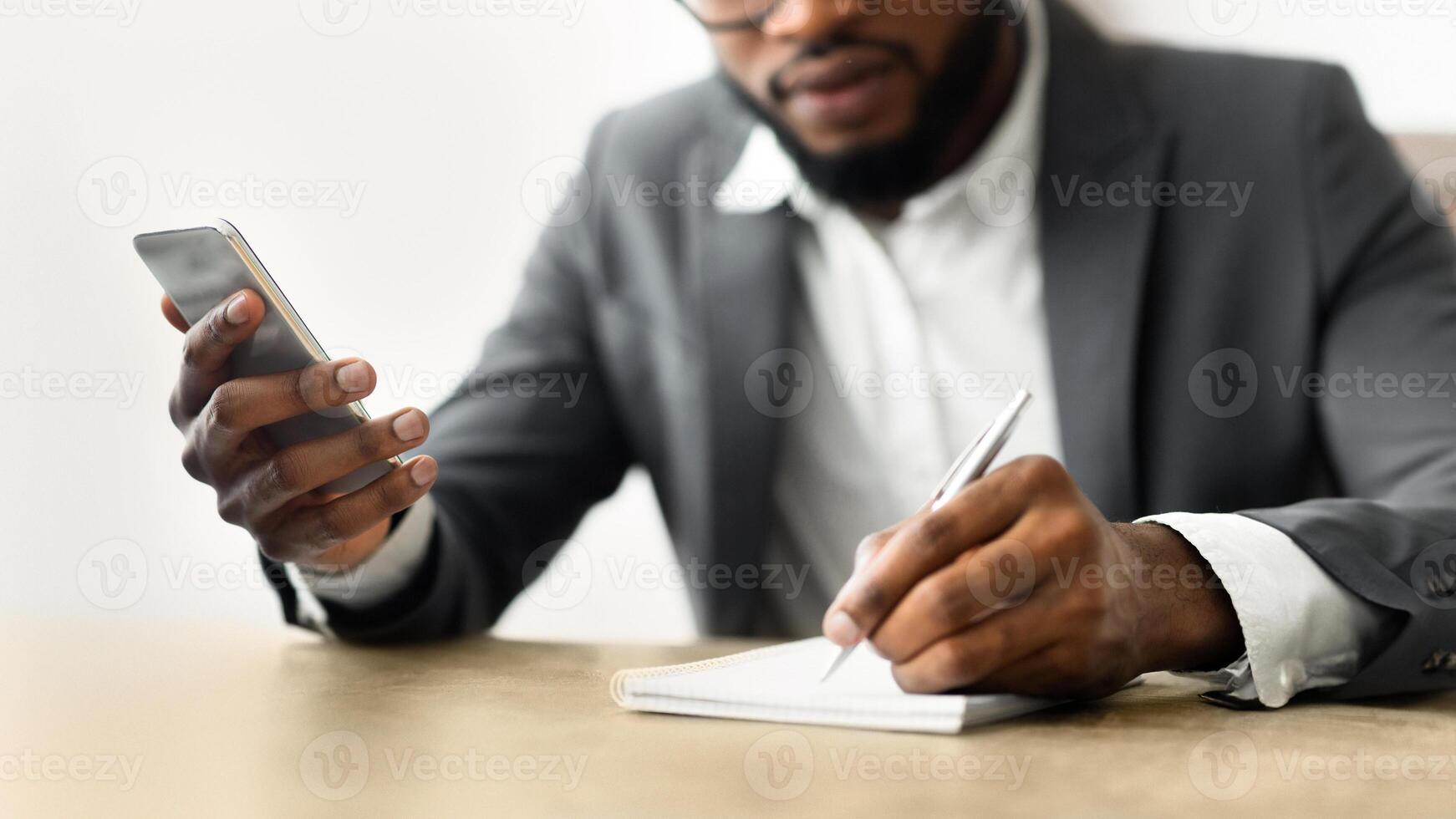A businessman is sitting at a desk, holding a phone in one hand and writing in a notebook with the other. photo