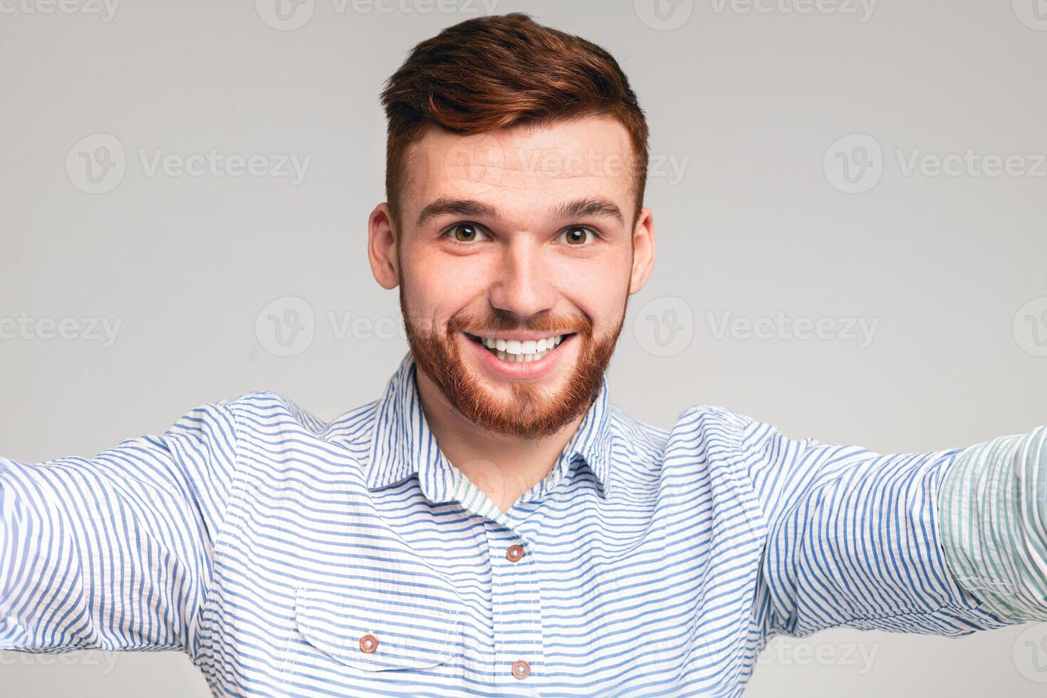 Young handsome bearded guy wearing casual shirt making selfie against beige studio background, cheerfully smiling photo