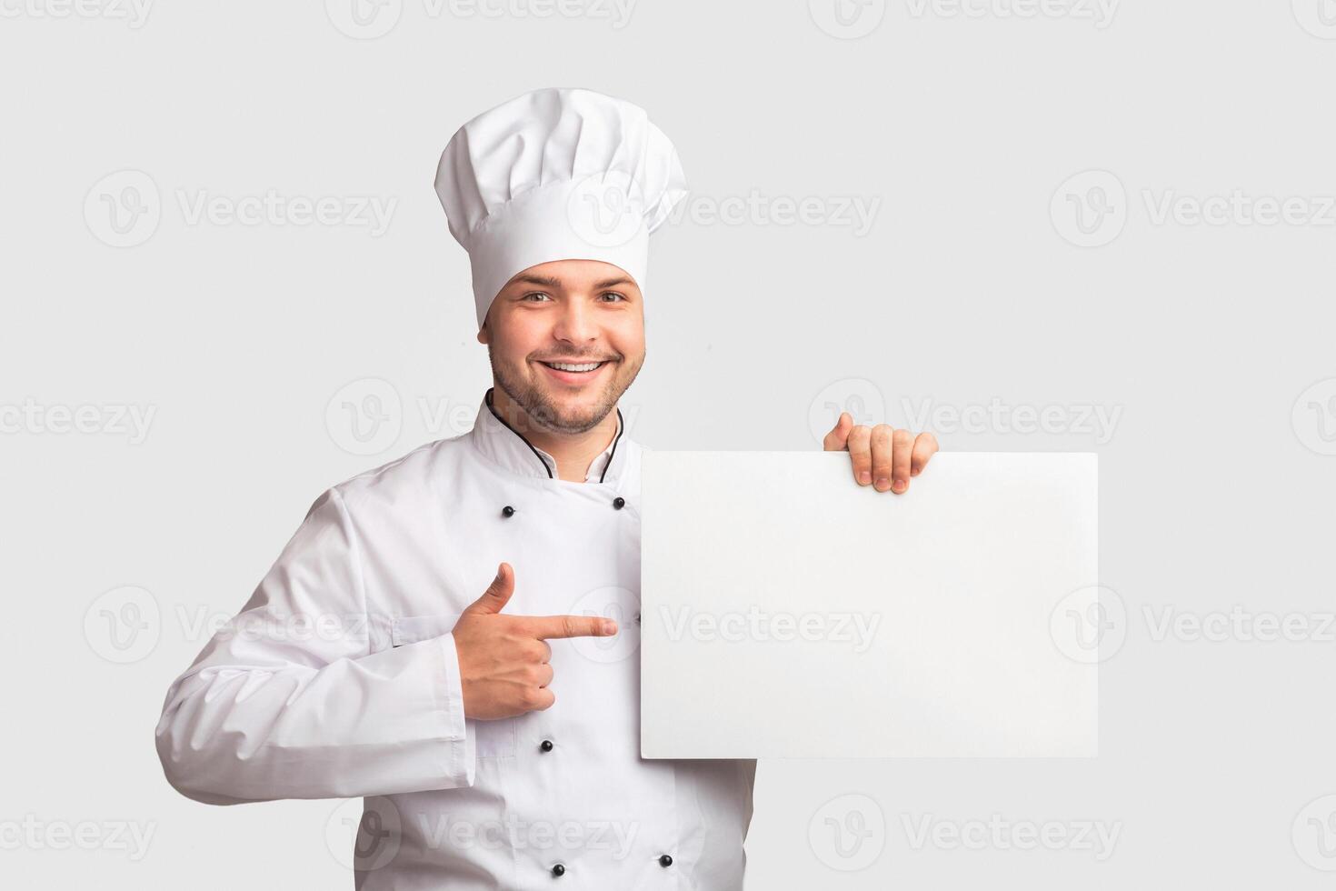 Chef Man Holding Blank White Paper Board Pointing Finger Recommending Best Recipes Standing Over Studio Background. Panorama, Mockup photo