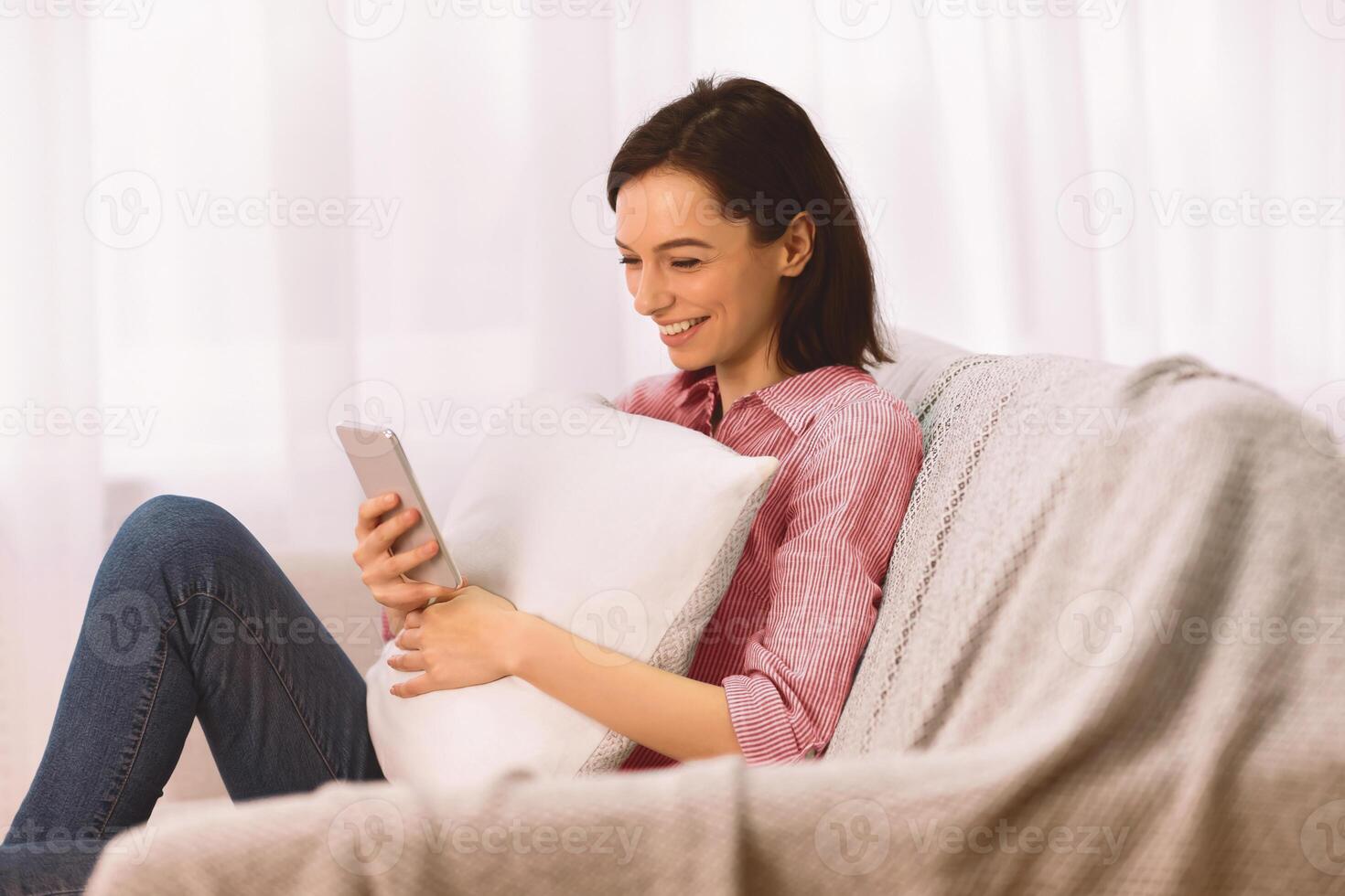 Free Time. Happy young woman holding pillow, sitting on sofa, using smartphone, copyspace photo