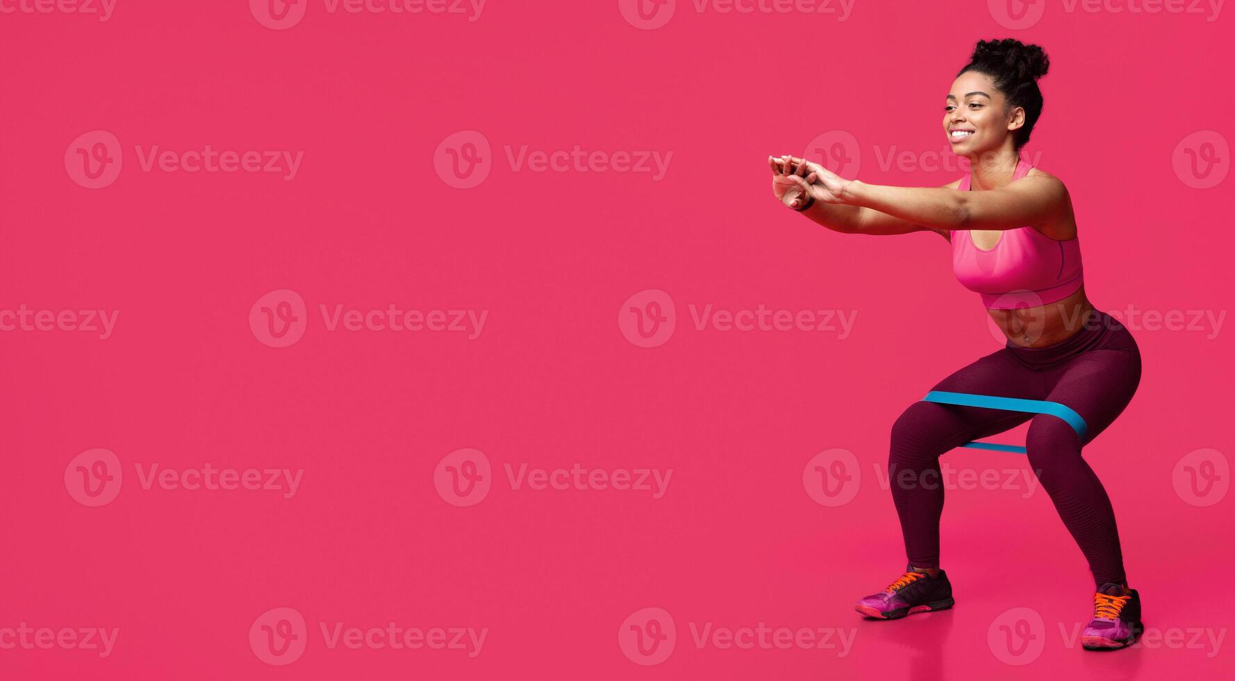 Young sporty smiling black woman doing stretching work out with elastic bands, squating over red studio background photo