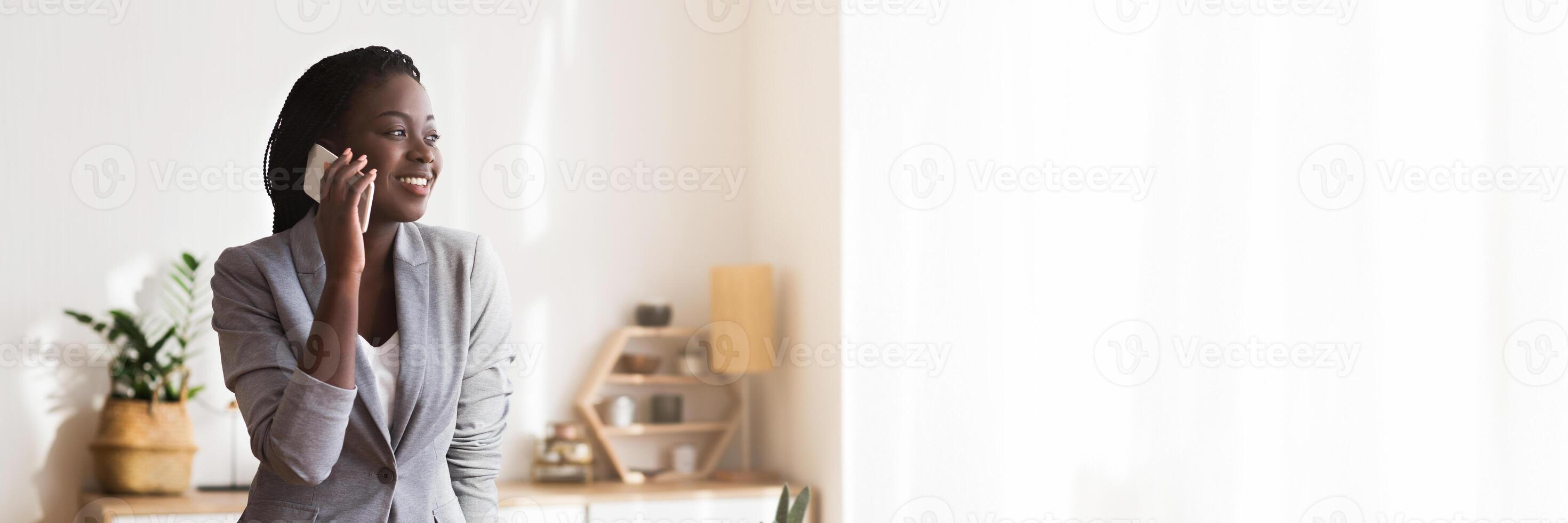 Business communication. Millennial black businesswoman talking on cellphone and leaning on office desk, free space photo